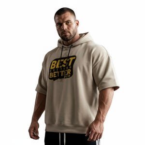 Könnte beinhalten: Beigefarbener Hoodie mit kurzen Ärmeln und dem Text "BEST GETTING BETTER" in Schwarz und Gelb. Der Hoodie hat eine Kapuze mit Kordelzug und eine lockere Passform. Das Bild ist vor einem weißen Hintergrund.