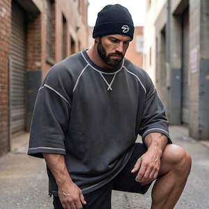 Peut inclure: Un bonnet noir avec un logo blanc et un sweat-shirt gris foncé avec des coutures blanches. Le sweat-shirt a des manches courtes et une coupe ample. La personne porte un short noir.