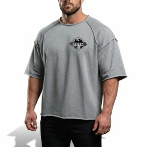 Pode incluir: Camiseta cinza de manga curta com gola redonda e bordas cruas. A camiseta apresenta um logotipo circular preto no peito com o texto "TETA" e "POWER & PERFORMANCE" em torno de um design de barra. Corte folgado.