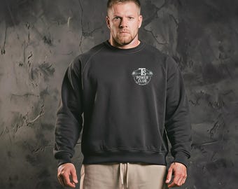 May include: A dark gray sweatshirt with the text "TS Power Club" in a white and gray design on the chest. The sweatshirt has a crew neck and long sleeves. The person in the image is wearing tan pants.