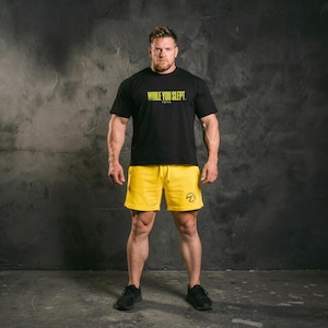 Könnte beinhalten: Eine Person trägt ein schwarzes T-Shirt mit dem gelben Text "WHILE YOU SLEPT", kombiniert mit gelben Shorts. Die Shorts haben einen Kordelzug und ein kleines Logo. Die Person trägt auch schwarze Sportschuhe.