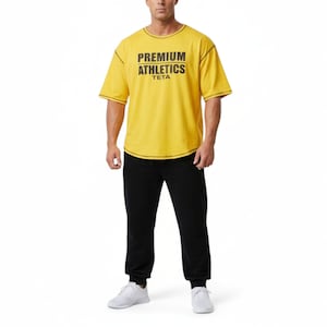Könnte beinhalten: Eine Person trägt ein gelbes T-Shirt und eine schwarze Jogginghose. Auf dem T-Shirt steht in schwarzer Schrift "PREMIUM ATHLETICS TETA". Die Jogginghose hat einen Kordelzug und elastische Bündchen. Weiße Turnschuhe vervollständigen das Outfit.