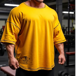 Könnte beinhalten: Senfgelbes, übergroßes T-Shirt mit schwarzen Nahtdetails. Das Shirt hat kurze Ärmel und einen runden Ausschnitt. Der Text "TS POWER CLUB WEAKNESS" ist auf der unteren rechten Seite des Shirts aufgedruckt.