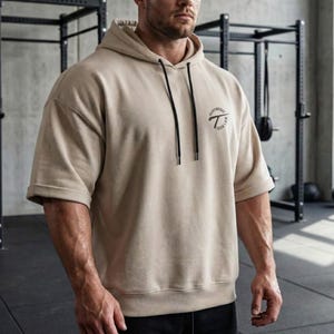 Könnte beinhalten: Beiger Hoodie mit kurzen Ärmeln und Kapuze mit Kordelzug. Der Hoodie hat ein schwarzes kreisförmiges Logo auf der Brust mit dem Text "HEAVYWEIGHT IRON LAW". Die Ärmel sind umgeschlagen. Im Hintergrund ist Fitnessgeräte zu sehen.