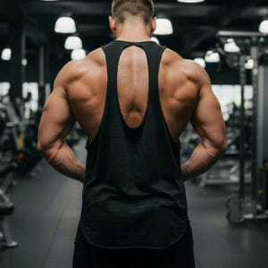 Könnte beinhalten: Ein schwarzes Tanktop mit Schlüsselloch-Rückenausschnitt, von hinten gesehen. Das Bild ist in einem Fitnessstudio aufgenommen, mit Trainingsgeräten im Hintergrund. Die Person, die das Tanktop trägt, hat gut definierte Muskeln.