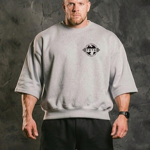 Pode incluir: Uma camisola cinza, grande, com mangas curtas. A camisola apresenta um logótipo preto no peito que diz "TETA" com o texto "POWER & PERFORMANCE BEYOND HUMAN LIMITS" à volta. A pessoa está a usar calções pretos e um relógio preto.