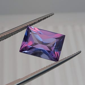 Peut inclure: Une pierre précieuse de coupe rectangulaire, avec un effet de changement de couleur du violet au bleu. La gemme est maintenue par une pince, révélant ses facettes. La pierre précieuse est un affichage vibrant de couleurs.