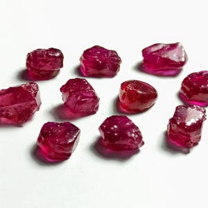 Könnte beinhalten: Eine Sammlung von grob geschnittenen, tiefroten Rubin-Edelsteinen. Die Steine haben unregelmäßige Formen und ein natürliches, unpoliertes Aussehen. Die Edelsteine sind auf einer weißen Oberfläche verstreut und zeigen ihre lebendige Farbe und Kristallstruktur.