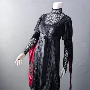 May include: Black vintage-style dress with intricate lace detailing on the bodice and sleeves. The dress features a high neckline, a black ribbon belt, and a red accent panel with black embroidery. The Last Kult is printed at the bottom.