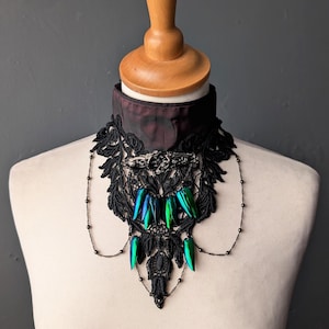 May include: A black lace choker featuring a dark red fabric band, an ornate silver-toned metal centerpiece, and dangling iridescent green beetle wing accents. The design includes delicate silver-toned chains draped across the front.