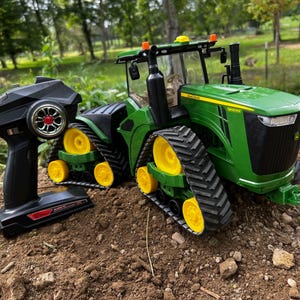 May include: A green and yellow toy tractor with a black remote control. The tractor has large black treads and yellow wheels. The remote control is black with a red and silver wheel.