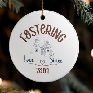 May include: White ceramic Christmas ornament with the words "Fostering Love Since 2001" in brown and navy blue. A house illustration is in the center. The ornament hangs from a gold string.