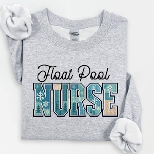 May include: A light grey sweatshirt featuring the text "Float Pool NURSE" in a winter design. The word "NURSE" is in block letters with snowflakes and fir trees. The sweatshirt has a crew neck and folded sleeves.