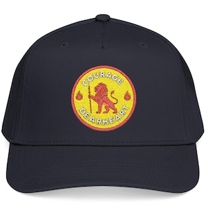 Op de afbeelding: Marineblauwe baseballcap met een geborduurde cirkelvormige patch. De patch toont een rode leeuw met een speer, met de woorden "COURAGE DEARHEART" in rood en geel. Twee gele bollen flankeren de leeuw.