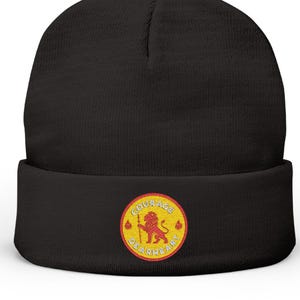 May include: Black beanie with a rolled cuff. A circular patch on the cuff features a yellow lion holding a spear, with the words "Courage Dearheart" in a circular pattern around the lion. The patch is outlined in red.