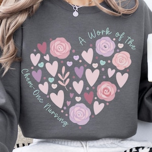 May include: Dark grey sweatshirt featuring a heart design made of pink and purple hearts, roses, and leaves. The text "A Work of the Chem-Onc Nursing" is printed on the sweatshirt. A white mug is held in the right hand.