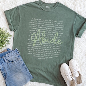 May include: Sage green t-shirt with the word "Abide" in a script font and a block of text. The shirt is paired with blue denim jeans and white sneakers. The text on the shirt is from John 15:9-17.