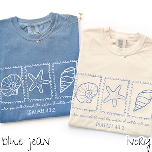 May include: Two t-shirts, one blue jean and one ivory, with a seashell, starfish, and conch design. The text "when you walk through the waters I will be with you ISAIAH 43:2" is printed below the design. Each shirt has a gold necklace.