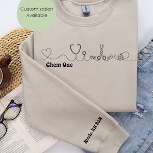 May include: A beige sweatshirt with a black line art design of a heart, stethoscope, scissors, and medical supplies. The text "Chem Onc" is below the design, and "Nicole. RN BSN" is on the sleeve. Customisation is available.