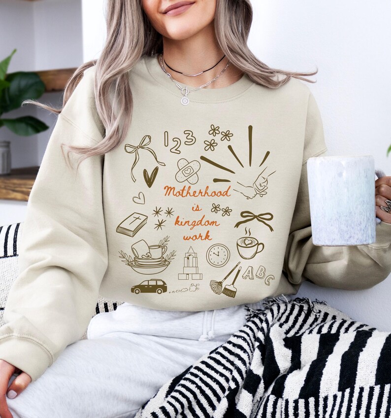 May include: Beige sweatshirt with brown graphic illustrations and the text "Motherhood is kingdom work." The design includes a car, coffee cup, clock, and other symbols. The sweatshirt is a neutral color and has a crew neck.