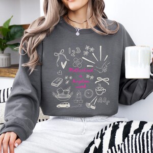 May include: A dark gray sweatshirt with a white doodle design featuring the words "Motherhood is a kingdom word" in pink. The design includes drawings of a bow, flowers, numbers, a bandage, a handshake, a book, a car, and a coffee cup.