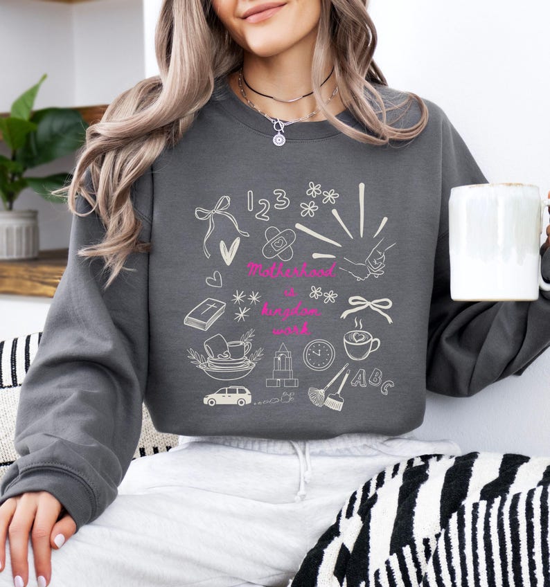May include: A dark gray sweatshirt with a white doodle design featuring the words "Motherhood is a kingdom word" in pink. The design includes drawings of a bow, flowers, numbers, a bandage, a handshake, a book, a car, and a coffee cup.