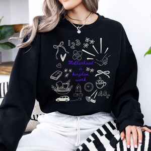 May include: Black sweatshirt with white and purple graphic illustrations. The design includes numbers, flowers, a bandage, a heart, a book, a coffee cup, a car, a clock, and the text "Motherhood is kingdom work."