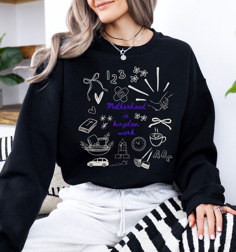 May include: Black sweatshirt with white and purple graphic illustrations. The design includes numbers, flowers, a bandage, a heart, a book, a coffee cup, a car, a clock, and the text "Motherhood is kingdom work."