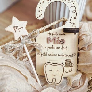 May include: Wooden tooth fairy door hanger with star and wand. The hanger has a star-shaped wand with the name "Mia" and a bow. The main sign has the text "Mia" and a tooth graphic. The hanger is decorated with stars.