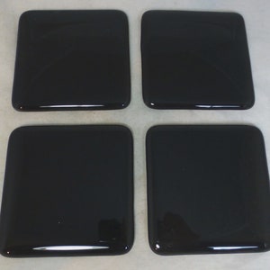 May include: Four square, glossy black coasters. Each coaster has rounded corners and a smooth, reflective surface. The coasters are arranged in a square formation, suitable for protecting surfaces from drink rings.
