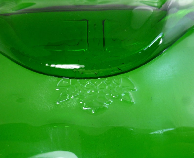 May include: Close-up of a green glass bottle with a textured surface. The bottle contains a clear liquid and has a raised design of a cluster of leaves on the bottom.