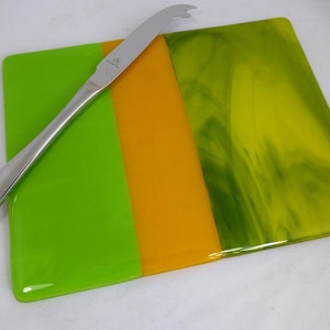 May include: A rectangular glass cheese board with a green, yellow and green striped design. A silver cheese knife is placed on the board.