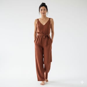 May include: A rust-colored jumpsuit with a V-neck and wide legs. The jumpsuit features a tie belt at the waist and pockets. The model is standing barefoot in front of a white background.