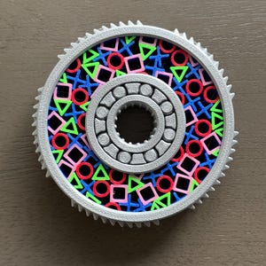 Könnte beinhalten: Ein graues, zahnradförmiges Objekt mit einem zentralen Lager und einem gemusterten Innenraum. Der Innenraum weist eine farbenfrohe Anordnung aus roten Kreisen, grünen Dreiecken, blauen Kreuzen und rosa Quadraten auf. Der äußere Rand hat ein gezahntes Design.