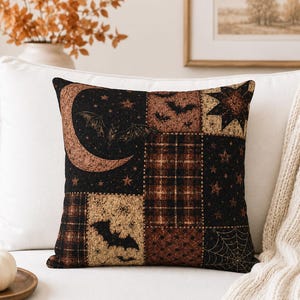 May include: A square throw cushion featuring a patchwork design in black, brown, and tan. The pattern includes illustrations of bats, a crescent moon, stars, a spider web, and plaid textures. The fabric has a woven appearance with a dark colour palette.