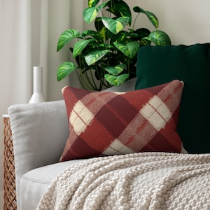 May include: A rectangular decorative pillow with a red and cream plaid pattern. The pillow is on a gray sofa with a cream-colored knitted throw. A green plant and a dark green pillow are in the background.