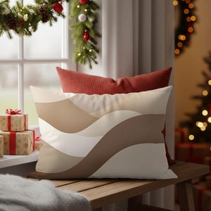Op de afbeelding: Een rechthoekig kussen met een golfpatroon in beige, bruin en wit. Het ligt op een houten bankje, met een rood kussen erachter. De scène bevat kerstversieringen bij een raam.