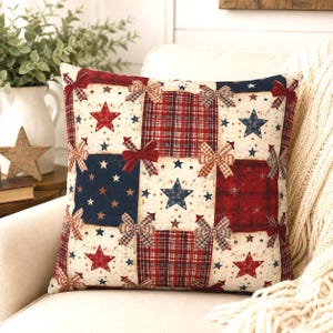 May include: Square decorative pillow with a patchwork design featuring red, white, and blue stars, bows, and plaid patterns. The pillow is on a white chair, suggesting a patriotic or Americana theme.