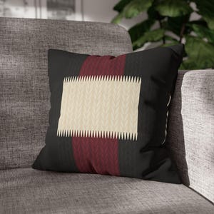 May include: Black cushion with a cream rectangular design featuring a leaf pattern. A vertical burgundy stripe and a black background complete the design. The cushion is placed on a grey sofa.