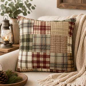 May include: A square throw pillow featuring a patchwork design of various plaid patterns in shades of deep red, forest green, cream, and tan. The fabric squares are stitched together to create a multi-colored grid across the entire surface.