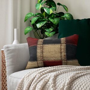 May include: A rectangular pillow with a plaid pattern in shades of red, black, and beige. The pillow is placed on a gray sofa, with a white blanket draped over it. A green plant and a dark green pillow are in the background.