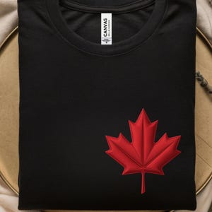 以下が含まれることがあります： 前面に赤い刺繍のメープルリーフが付いた黒いTシャツ。 シャツはきれいに折りたたまれ、クルーネックです。 ブランドラベル「Canvas Bella+Canvas」が右上に見えます。