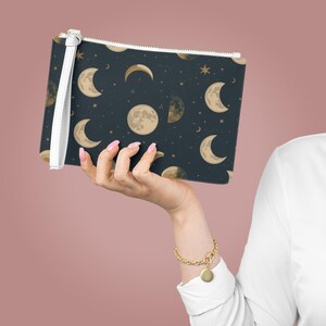 May include: A rectangular clutch bag with a navy blue background featuring a celestial moon phase pattern in gold. The bag has a white wrist strap and a gold zip. The accessory is held by a person.