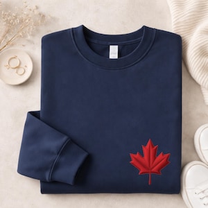 May include: A navy blue crewneck sweatshirt featuring a red maple leaf embroidered on the lower right side. The garment has long sleeves with ribbed cuffs and a ribbed neckline, showcasing a clean, solid-coloured design with a single focal point.