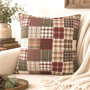 Peut inclure: Un coussin carré au motif patchwork composé de divers carrés de tissu à carreaux et écossais dans des tons de rouge profond, vert forêt, marron et crème. Le coussin présente un aspect structuré et rembourré avec des coutures visibles.