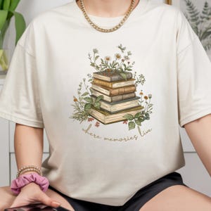 May include: Cream-colored t-shirt featuring a stack of books adorned with floral illustrations. The design includes daisies, greenery, and the text "where memories live" in a script font. The shirt has short sleeves.