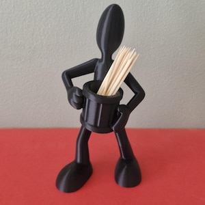 Puede incluir: Un palillero negro impreso en 3D con forma de figura estilizada. La figura sostiene un recipiente cilíndrico lleno de palillos de madera. La figura tiene un diseño minimalista con rasgos redondeados.