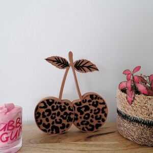 Peut inclure: Décoration en forme de cerises en bois avec un motif léopard. Les cerises sont marron clair avec des taches noires, et les feuilles sont détaillées avec des lignes noires. Posé sur une surface en bois.