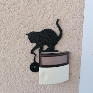 Può includere: Silhouette nera di gatto in metallo che gioca con un gomitolo di lana, montata sopra un campanello bianco e marrone. Il gatto è attaccato al campanello su un muro beige strutturato. La coda del gatto è curva verso l'alto.