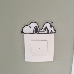 May include: A Snoopy-themed light switch cover. The cover features a white Snoopy figure with black outlines, lying on top of a white light switch. The background is a light green wall.